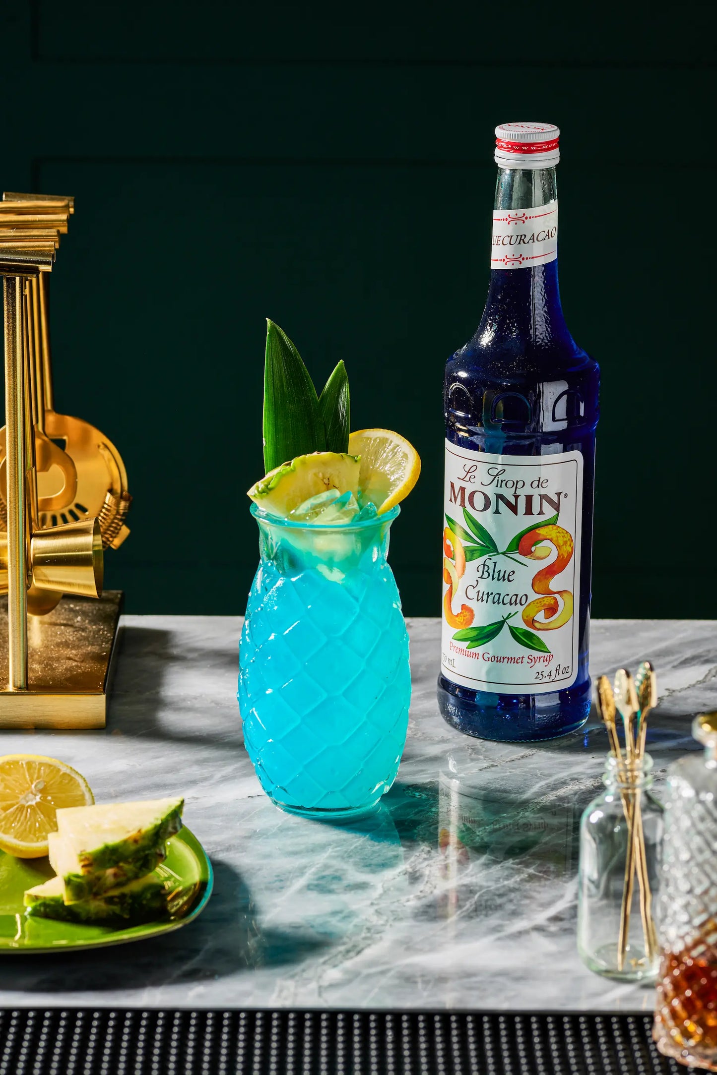 Monin Blue Curacao Syrup bottle beside a tropical blue cocktail garnished with pineapple and lemon