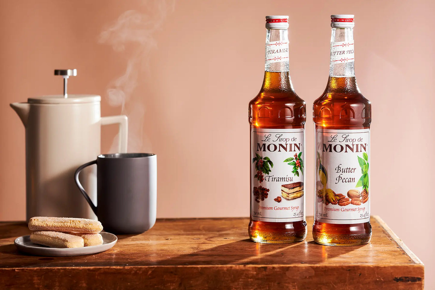 Two bottles of Monin syrup, one Butter Pecan and one Tiramisu, displayed on a wooden table next to a coffee pot.