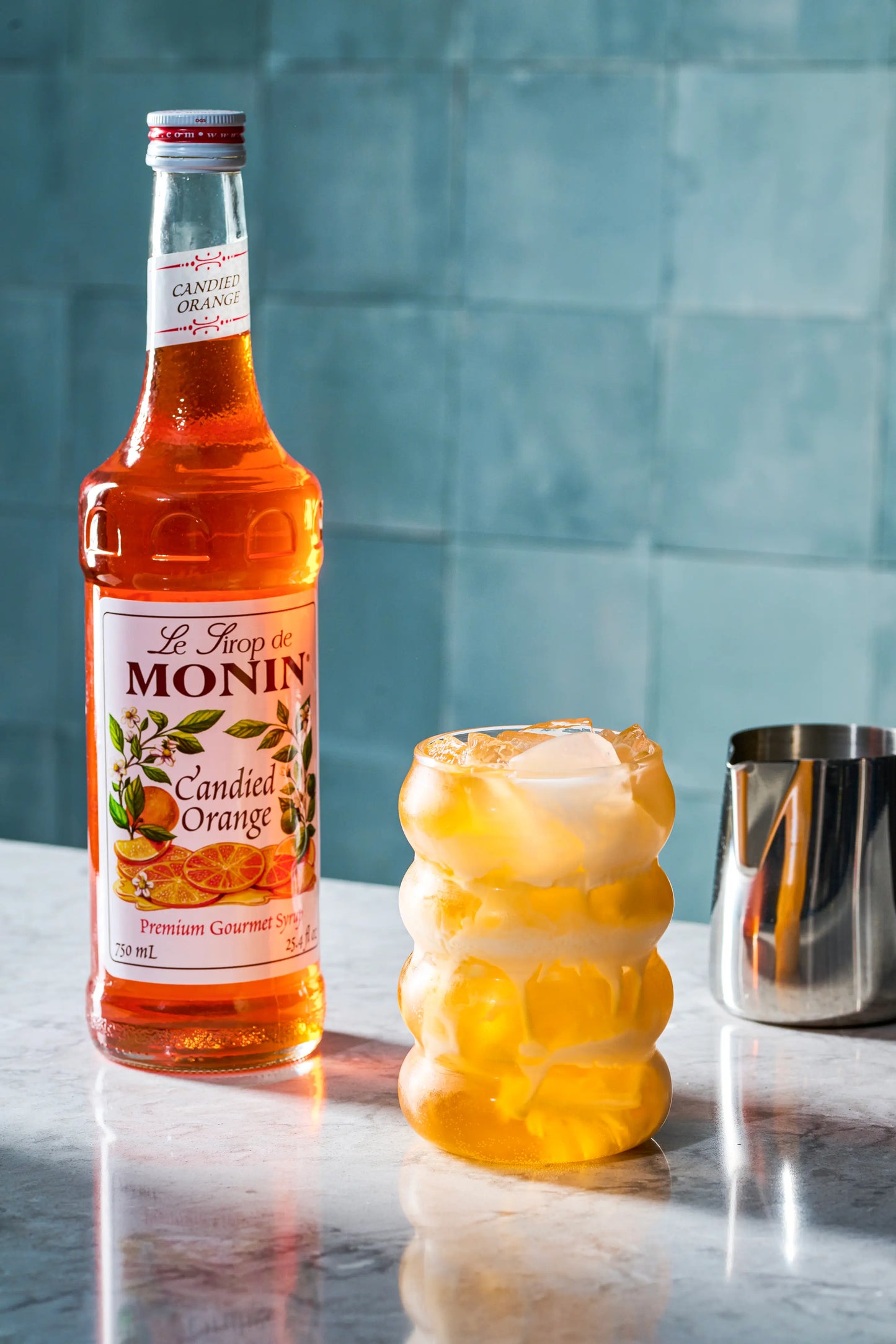Monin Candied Orange Syrup beside an iced orange beverage with creamy foam, showcasing bright citrus flavour for cocktails, mocktails and specialty drinks.