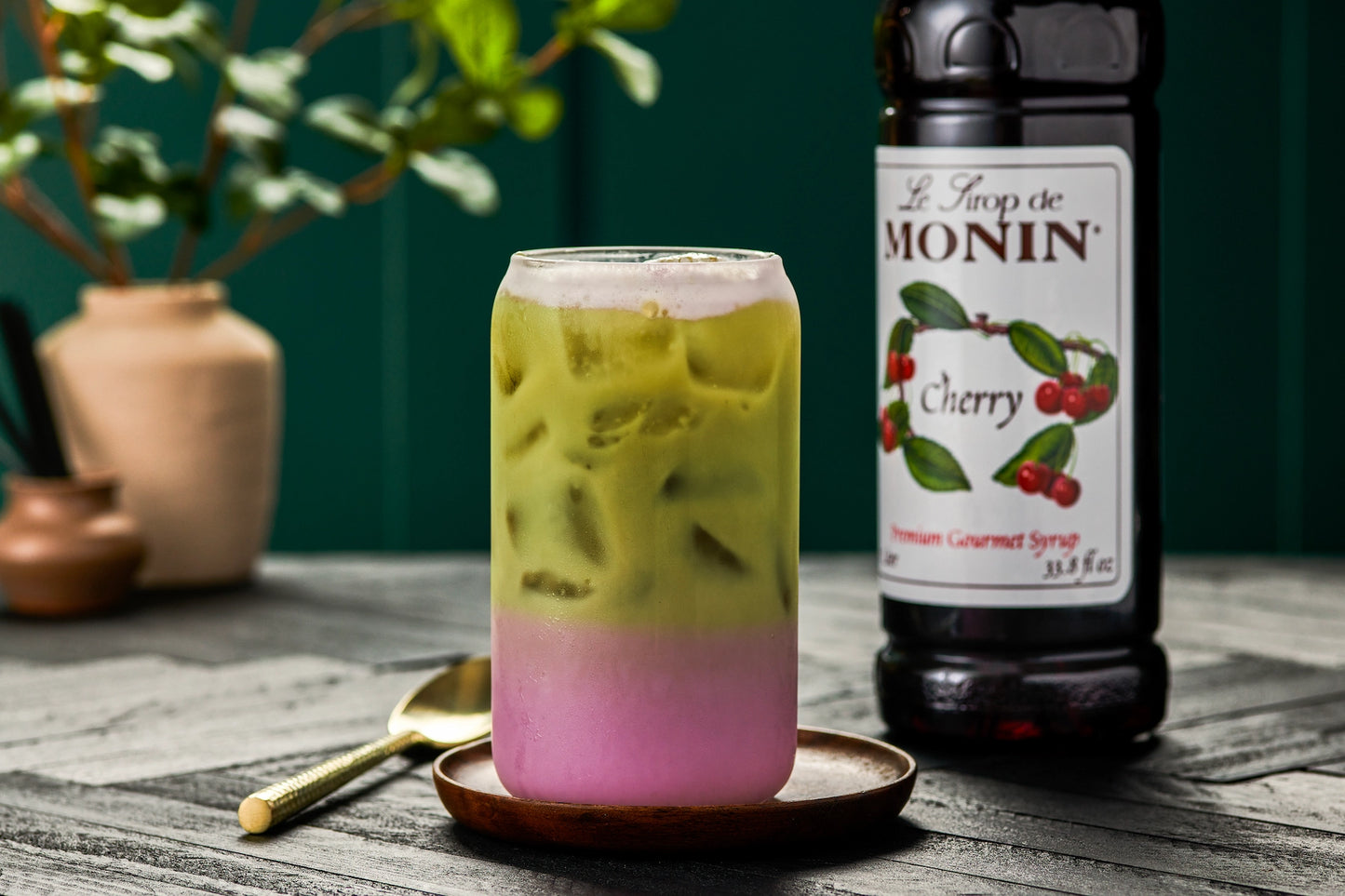 Monin Cherry Syrup beside a layered iced matcha latte with cherry-flavoured milk, showcasing a colourful cherry matcha drink.