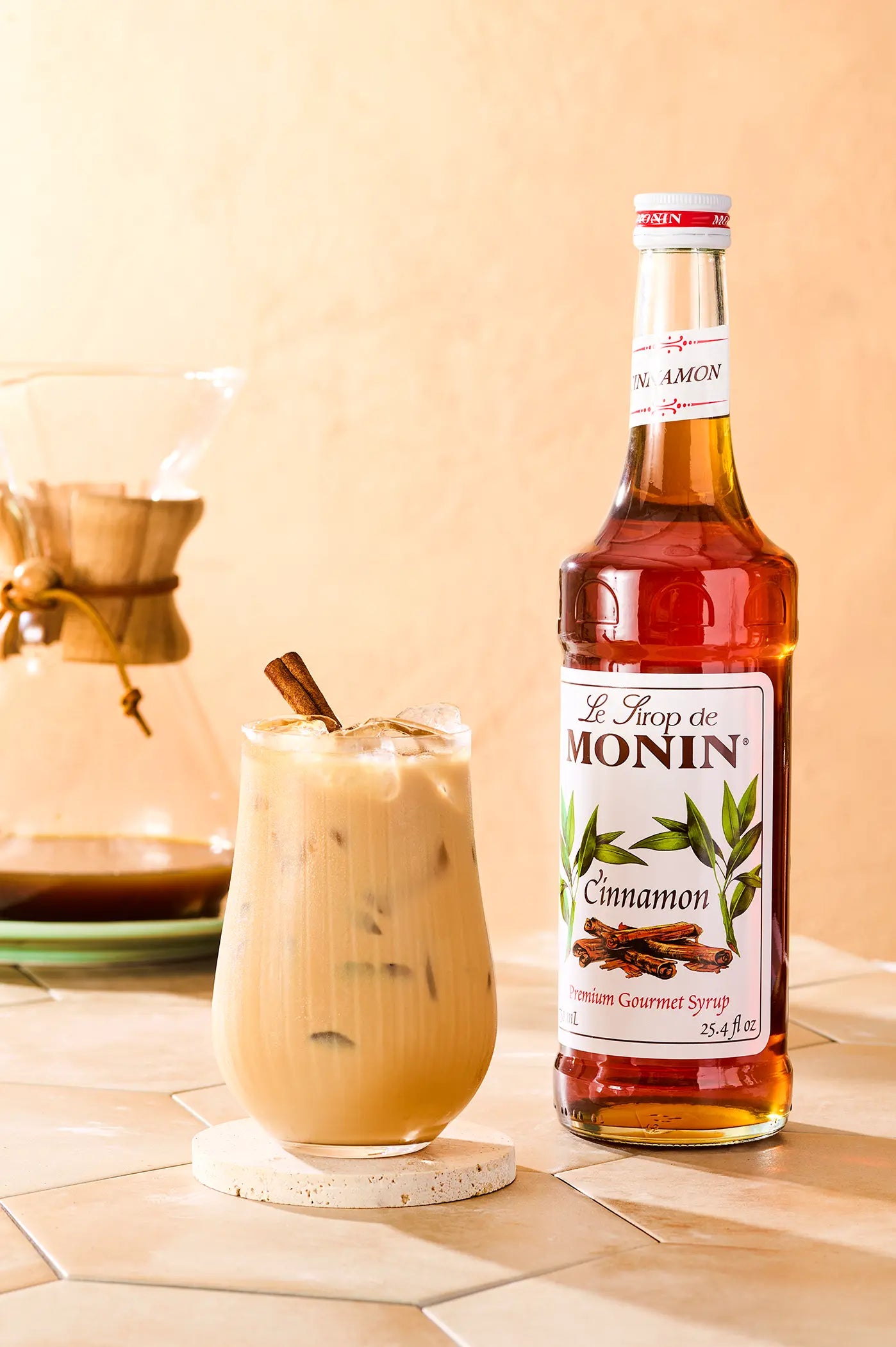Monin Cinnamon Syrup beside an iced cinnamon coffee garnished with a cinnamon stick, showcasing warm cinnamon flavour for specialty coffee drinks.