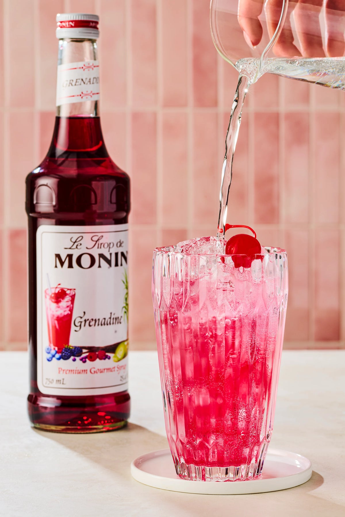 Monin Grenadine Syrup beside a bright red sparkling drink garnished with a cherry