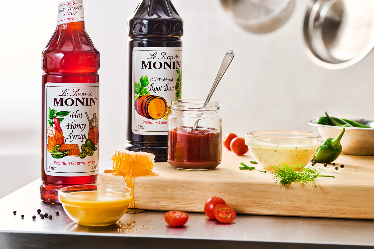 Monin Old Fashioned Root Beer Syrup with Hot Honey Syrup, sauces, tomatoes, peppers and honey on a kitchen counter.