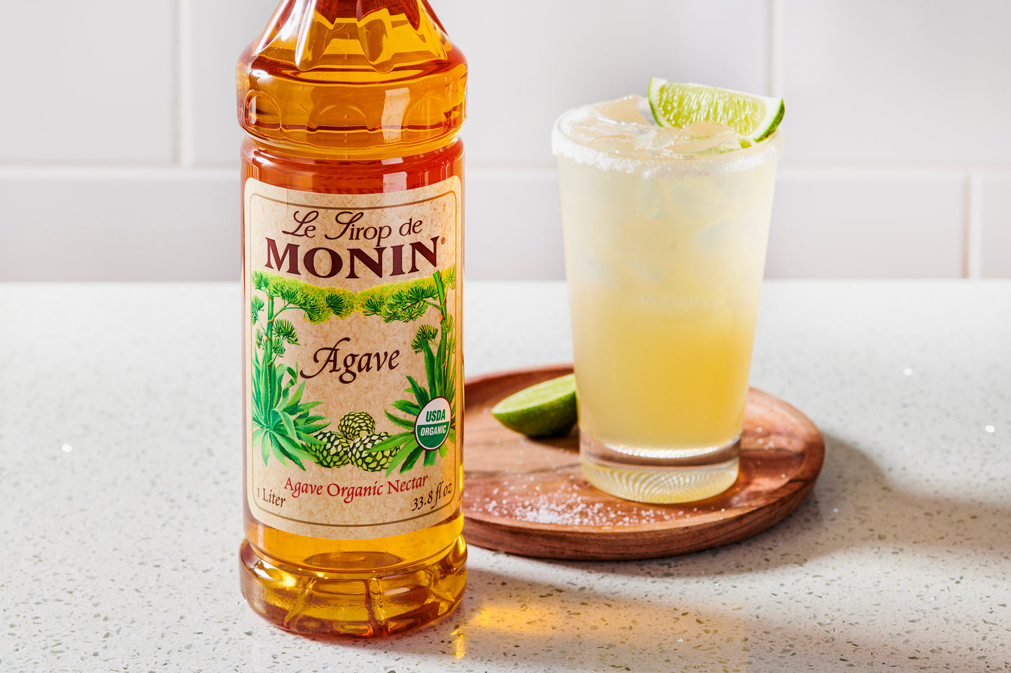 A bottle of Monin Agave organic nectar stands on a light countertop beside a margarita glass garnished with lime, on a round wooden coaster.