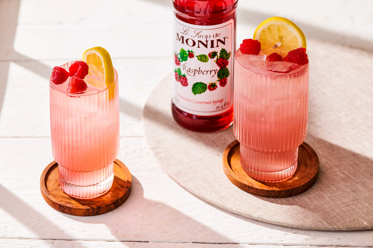 Monin Raspberry Syrup bottle beside refreshing raspberry lemonade garnished with fresh raspberries and lemon
