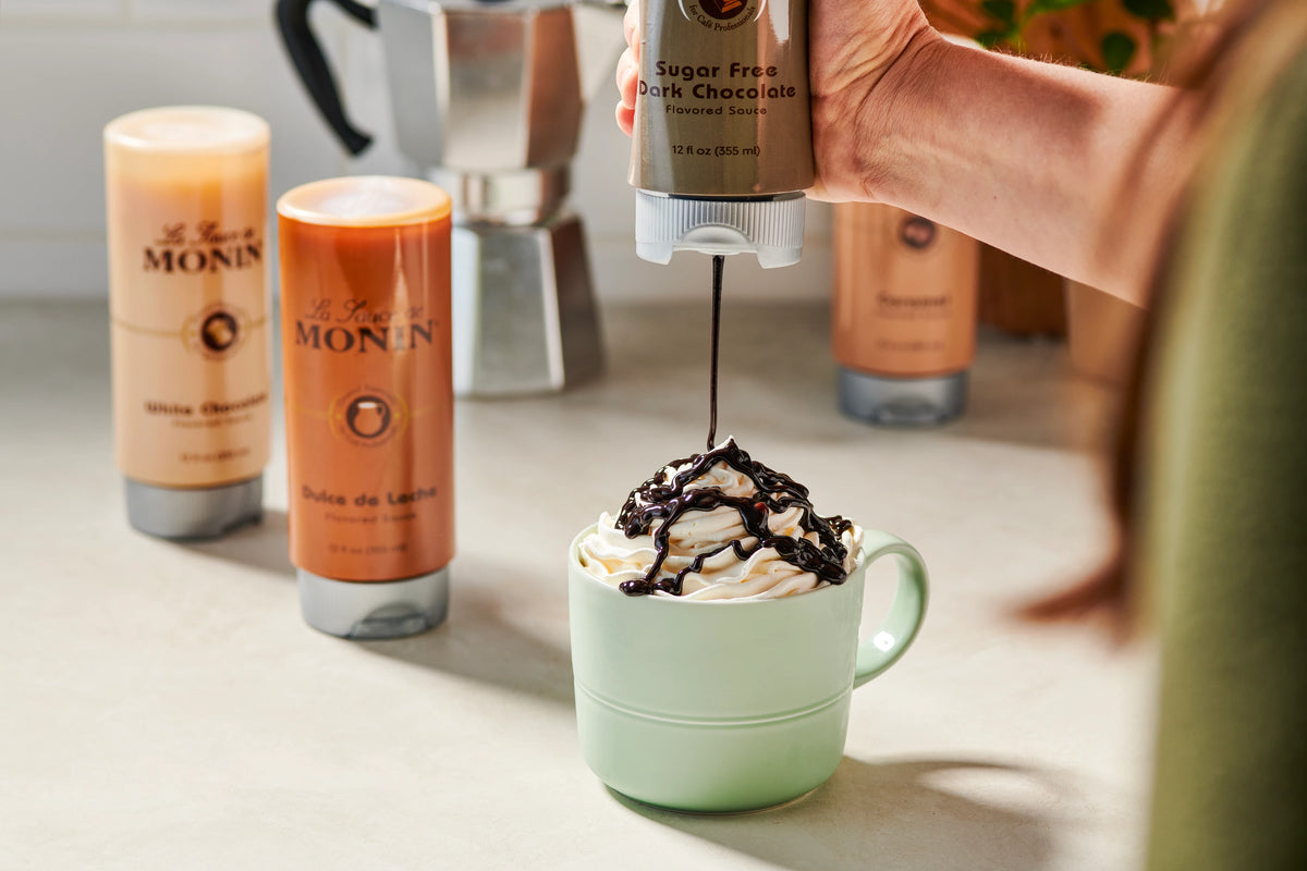 Monin Sugar Free Dark Chocolate Sauce drizzled over whipped cream on a hot chocolate, with other Monin sauces in the background
