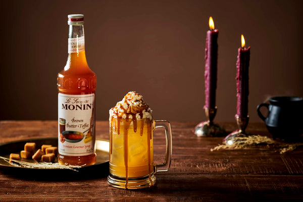 Iced caramel drink topped with whipped cream and toffee drizzle in a glass mug, alongside a bottle of Monin Brown Butter Toffee Syrup and lit candles.