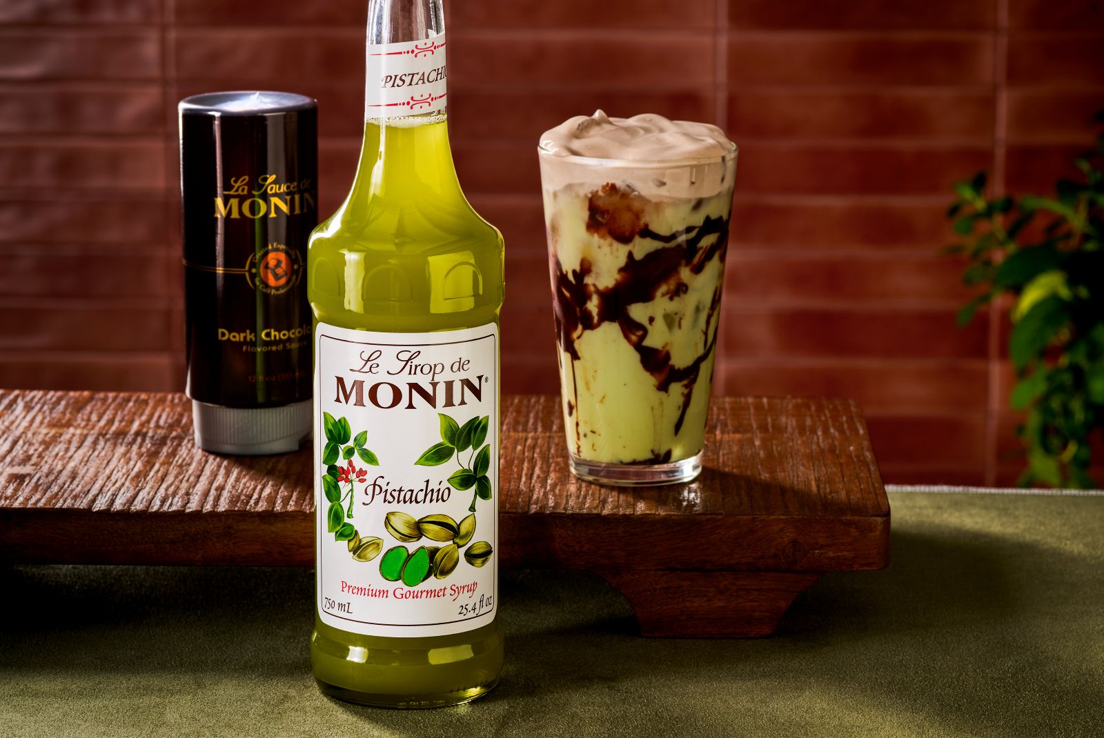 A bottle of Monin Pistachio Syrup in the foreground. A bottle of Monin Dark Chocolate Sauce and an iced Dubai Chocolate Matcha Latte in the background.