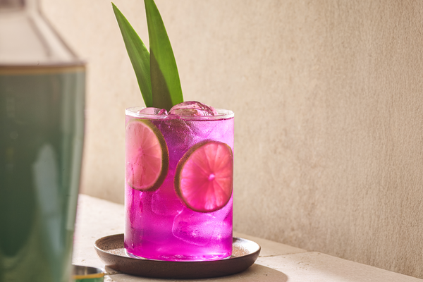 A bright purple Desert Pear mocktail in a rocks glass, garnished with lime wheels and pineapple leaves.