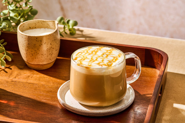 A caramel-drizzled latte in a clear glass mug on a wooden tray, styled with a ceramic milk pitcher and greenery in the background.