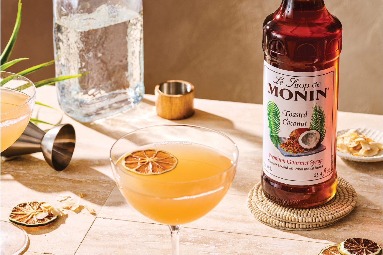 Monin Toasted Coconut Syrup bottle beside a tropical cocktail garnished with dried citrus and cocktail-making accessories