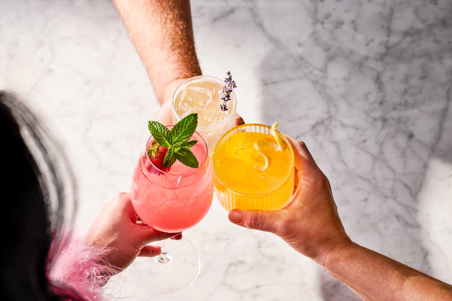 three hands holding colorful mocktails against a marble background