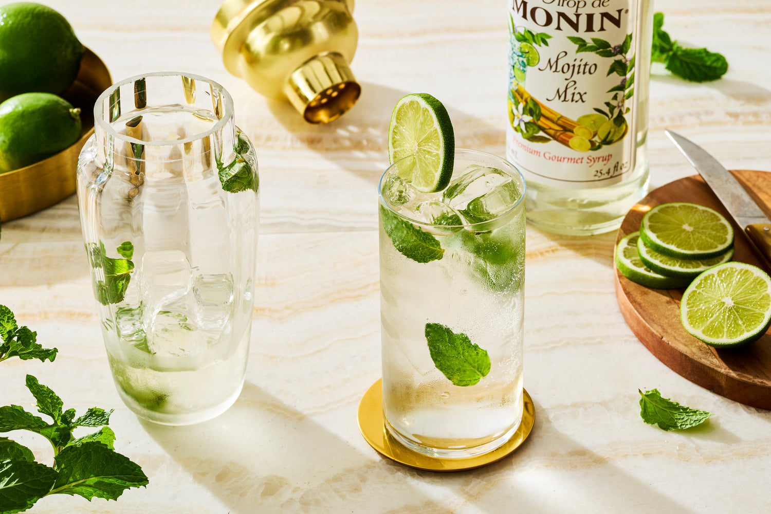 Two glasses of non-alcoholic mojito with lime slices and mint leaves on a marble surface with Monin Mojito Mix bottle.