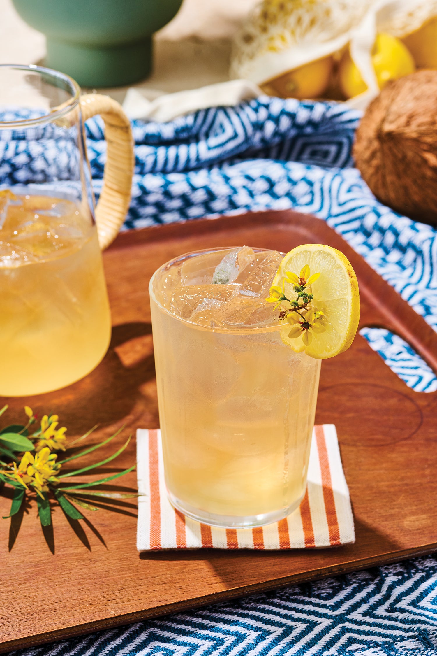 Glass of lemonade with a lemon slice on a wooden tray with a pitcher in the background.