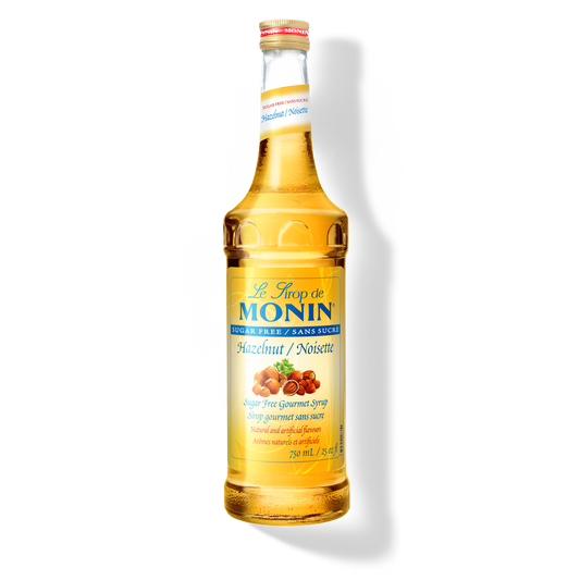 A glass bottle of Monin Sugar Free Hazelnut Syrup, with a yellow label displaying the product name and an image of hazelnuts.