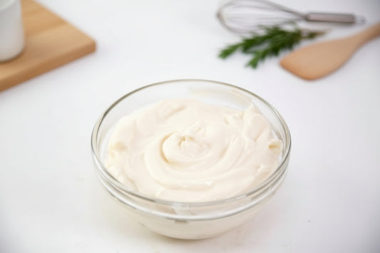 Glass bowl of white cream on a light surface with a whisk and rosemary in the background
