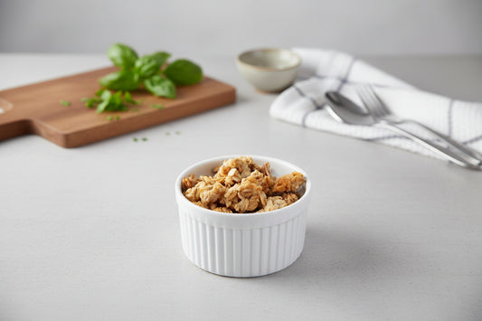 White ramekin with granola on a gray surface with basil and a towel in the background