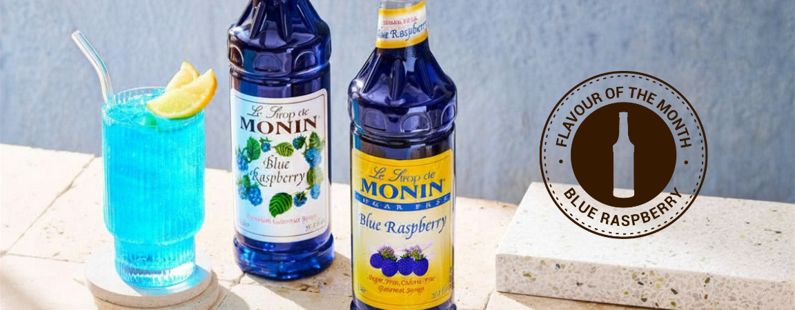 Two bottles of Monin Blue Raspberry syrup displayed next to a blue cocktail glass with a lemon slice, on a stone surface with a circular 'Flavor of the Month' badge.
