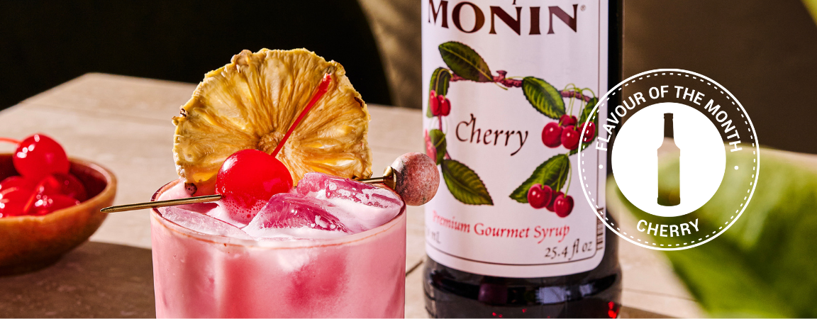 Monin Cherry Syrup bottle beside a cherry cocktail garnished with dried pineapple and cherry, featuring a Cherry Flavour of the Month badge