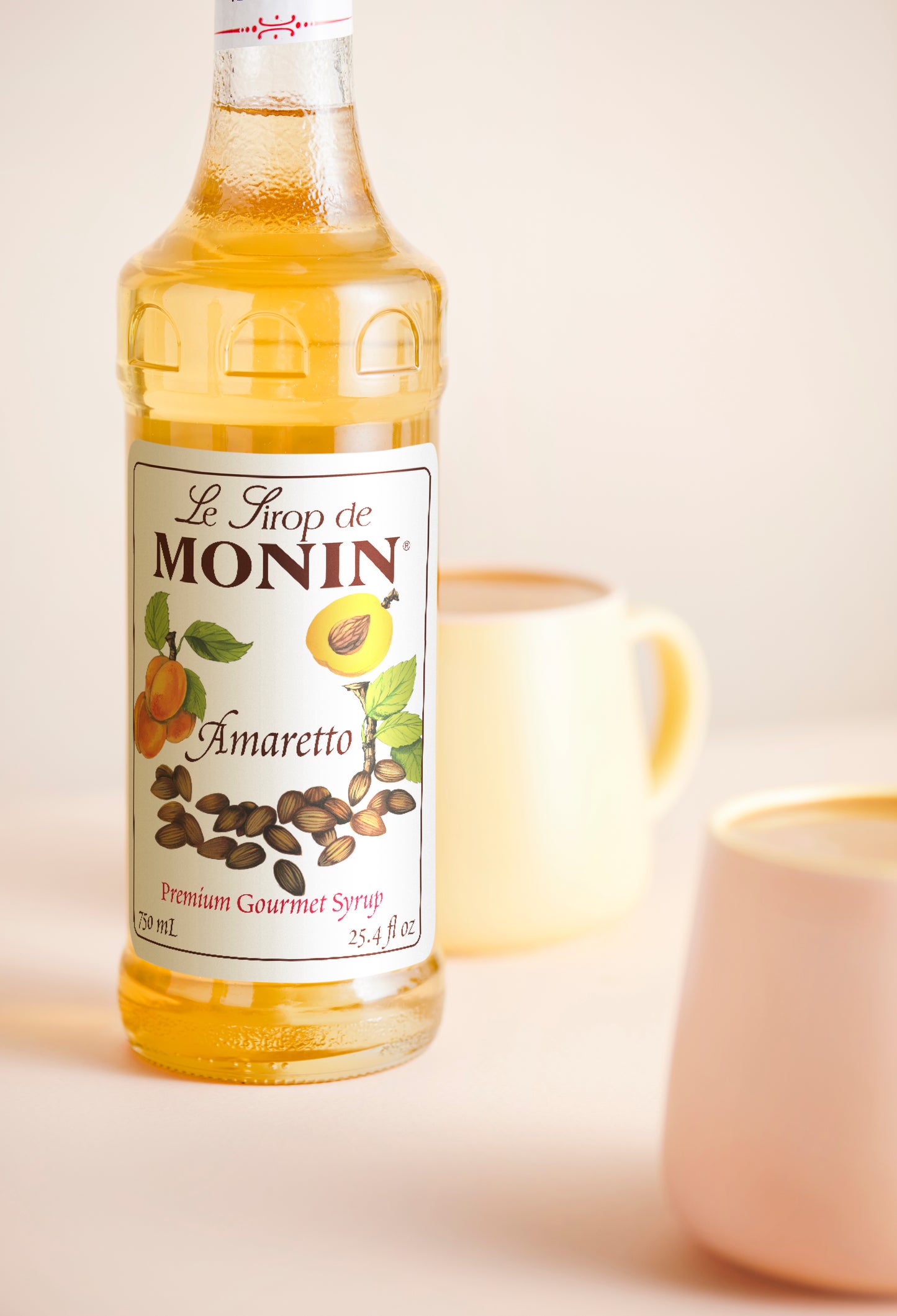 A bottle of Monin Amaretto syrup stands on a light beige surface with two frothy coffee cups blurred in the background.