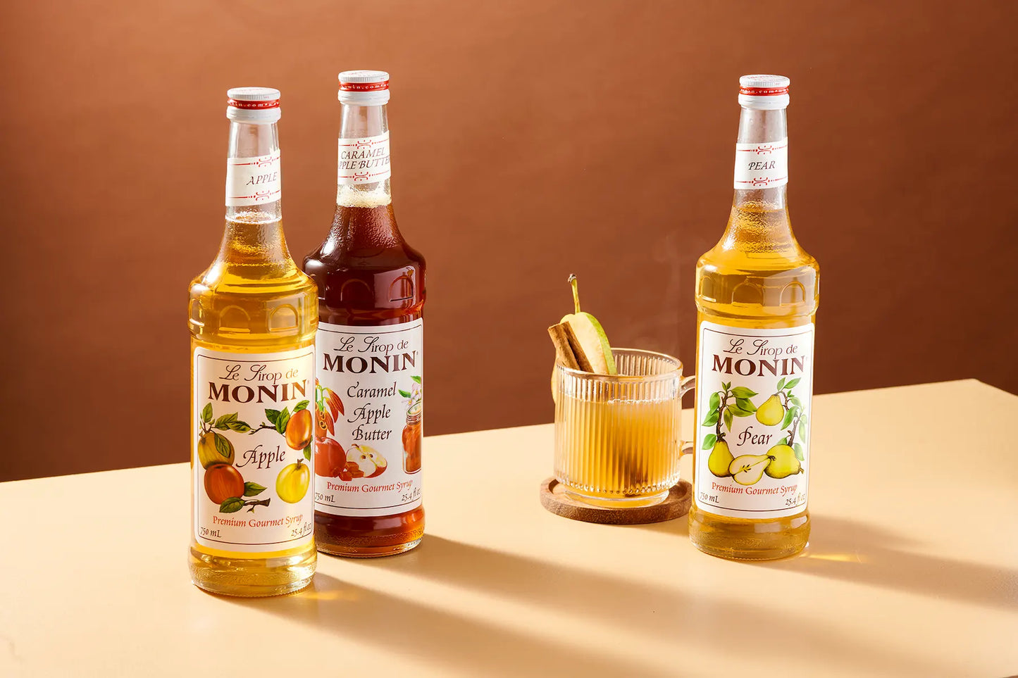 Three bottles of Monin syrup and a glass of mixed drink on a beige tabletop against a warm brown background. The bottles include Apple, Caramel Apple Butter, and Pear flavors.