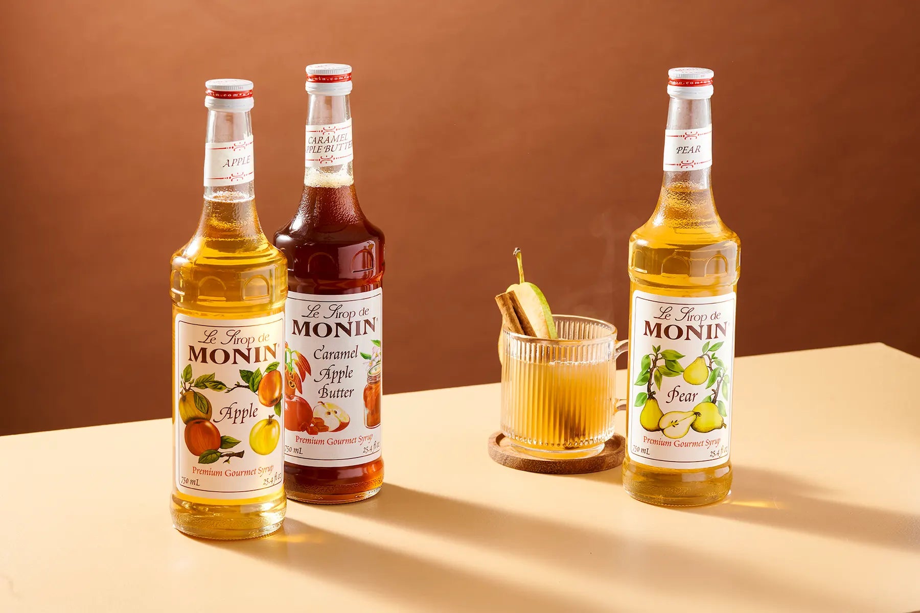Three bottles of Monin syrup and a glass of mixed drink on a beige tabletop against a warm brown background. The bottles include Apple, Caramel Apple Butter, and Pear flavors.