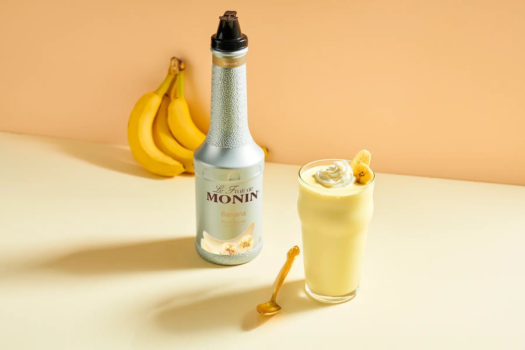 A bottle of Monin Banana puree with a black pourer cap stands next to a glass of pale-yellow banana drink topped with whipped cream and a banana slice, with bananas and a spoon on a beige surface. 