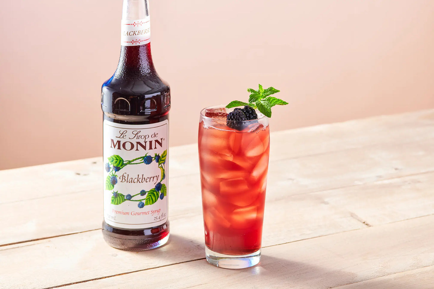 A bottle of Monin Blackberry syrup stands next to a tall glass of red iced cocktail with a blackberry and mint garnish on a light wooden table.