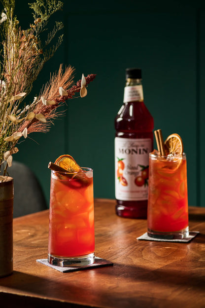 Two tall glasses of orange-red iced drink garnished with orange slices, set on a wooden table beside a bottle of Monin Blood Orange syrup, with dried flowers in the background.
