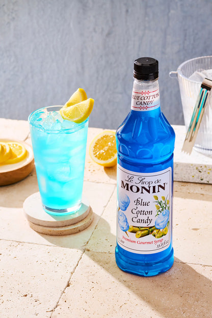 A bottle of Monin Blue Cotton Candy syrup stands on a stone surface beside a glass of bright blue cocktail with lemon slices.