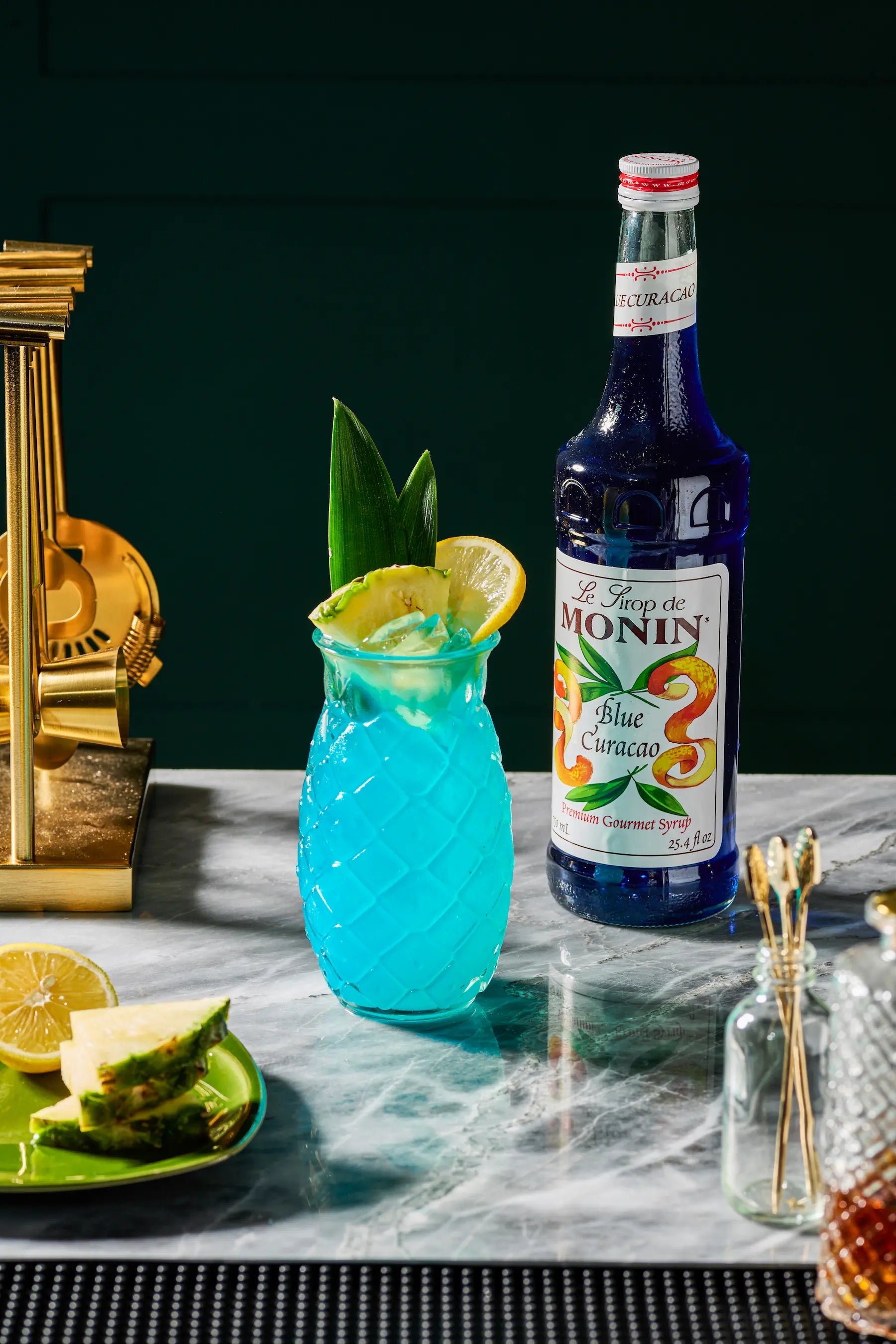 Monin Blue Curacao Syrup bottle beside a tropical blue cocktail garnished with pineapple and lemon