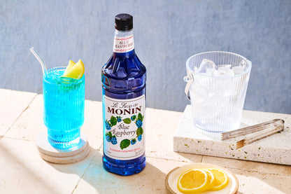 Monin Blue Raspberry Syrup bottle beside a bright blue raspberry drink garnished with lemon