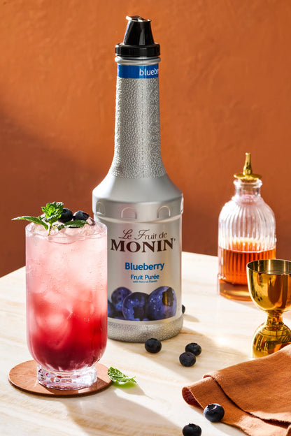 Monin Blueberry Purée bottle beside a refreshing blueberry drink with ice, fresh blueberries and mint