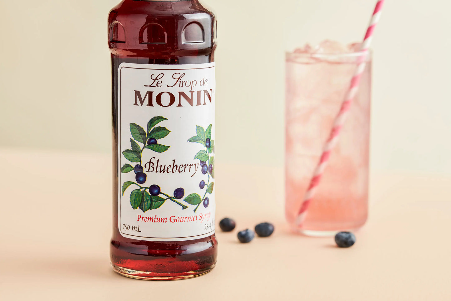 Monin Blueberry Syrup bottle beside a refreshing blueberry drink with ice, fresh blueberries and a striped straw