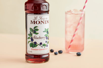 Monin Blueberry Syrup bottle beside a refreshing blueberry drink with ice, fresh blueberries and a striped straw