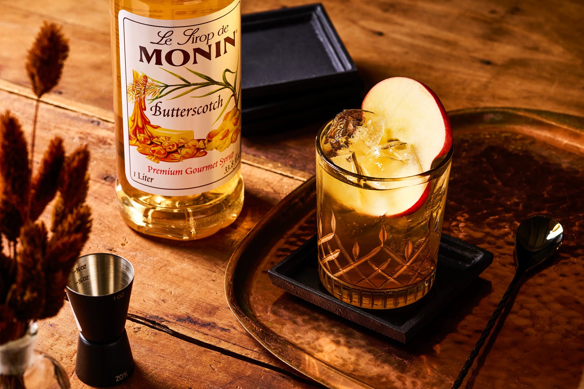 Monin Butterscotch Syrup beside a butterscotch apple cocktail served over ice and garnished with a fresh apple slice.