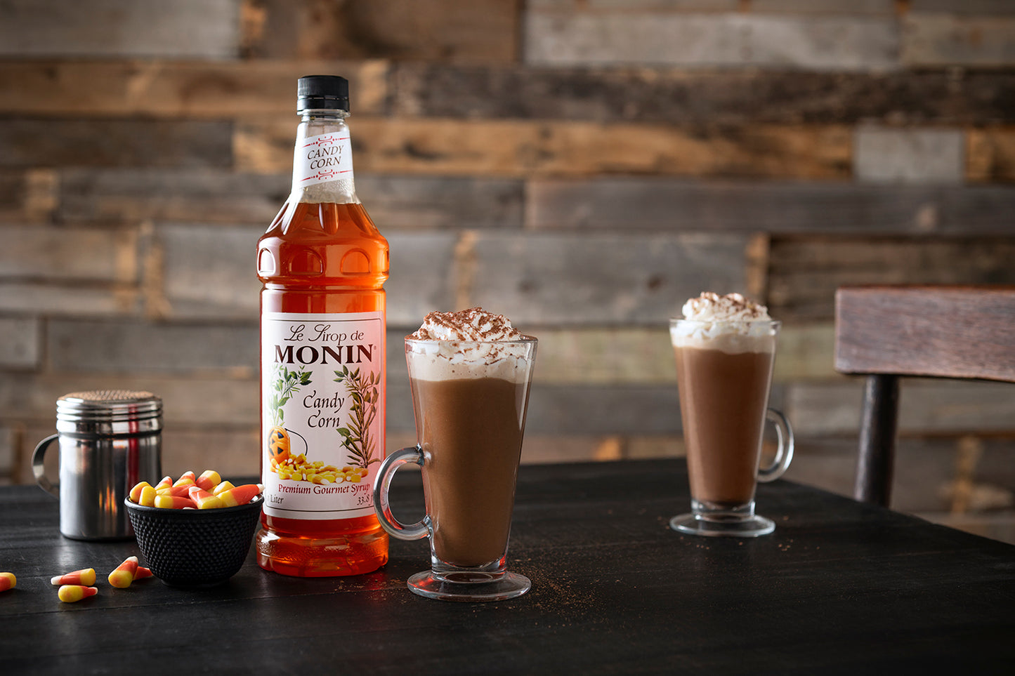Monin Candy Corn Syrup beside hot chocolate topped with whipped cream, with candy corn for a festive fall and Halloween beverage.