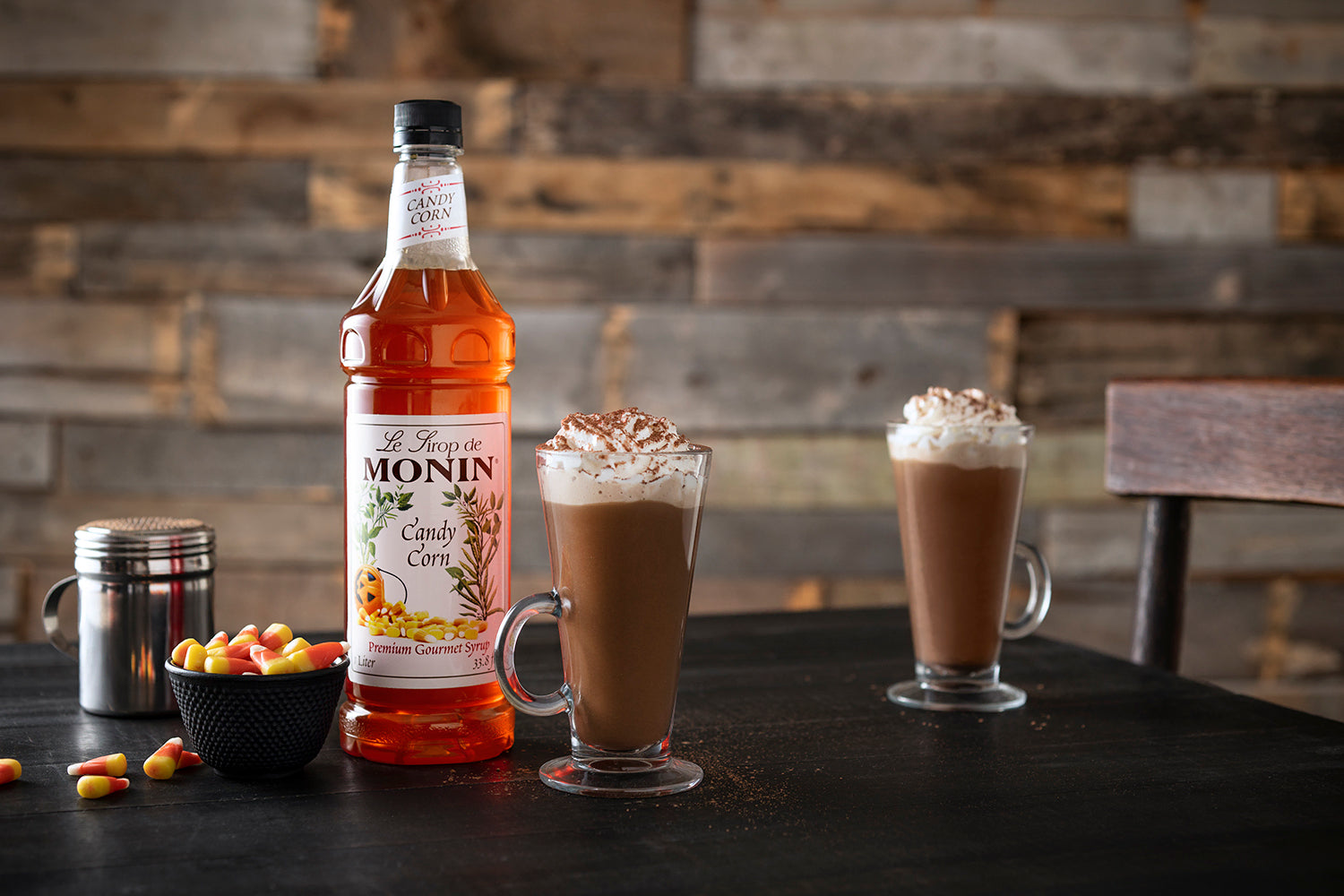 Monin Candy Corn Syrup beside hot chocolate topped with whipped cream, with candy corn for a festive fall and Halloween beverage.