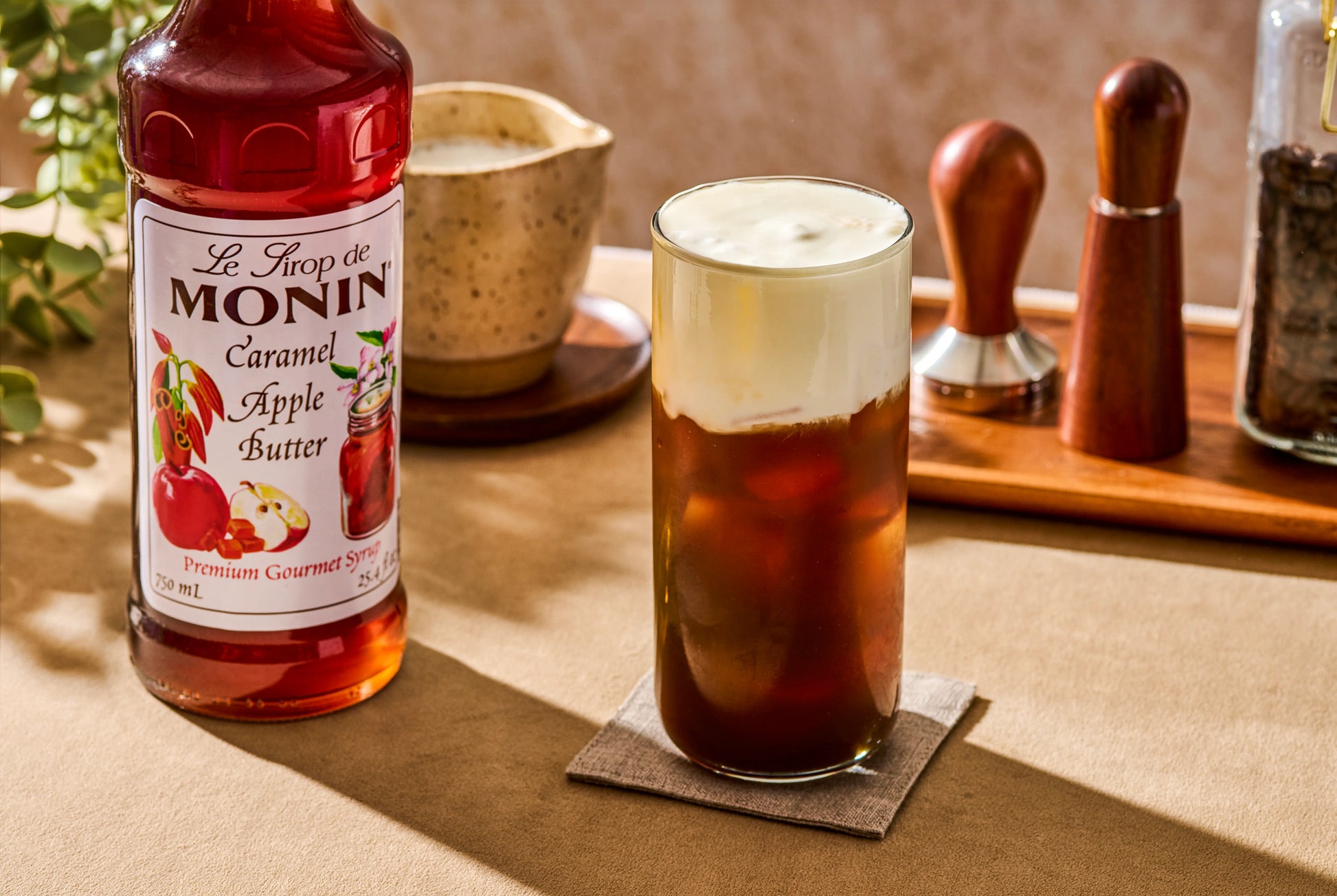 Monin Caramel Apple Butter Syrup beside an iced coffee topped with creamy cold foam, featuring rich caramel apple flavour for a seasonal fall drink.