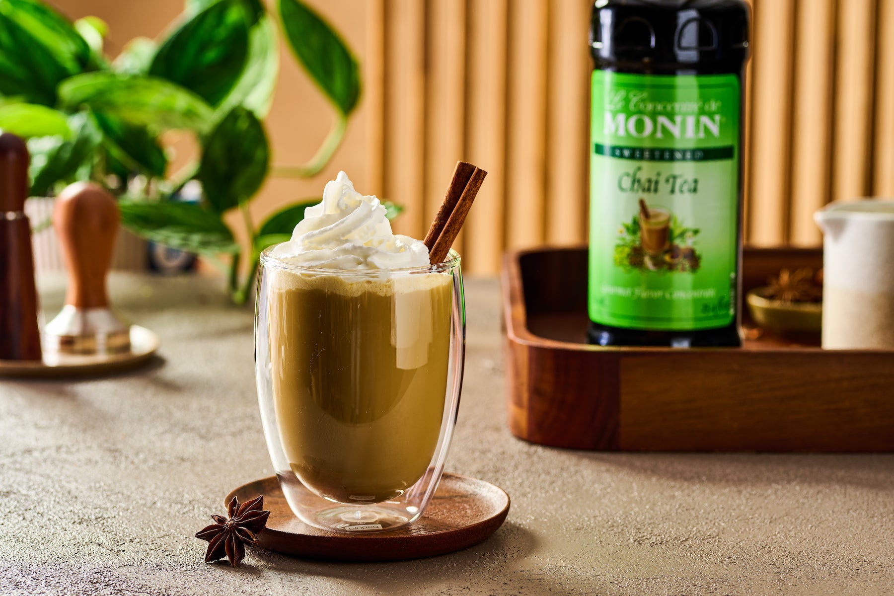 Monin Chai Tea Concentrate beside a creamy chai latte topped with whipped cream and a cinnamon stick, featuring warm chai spices.