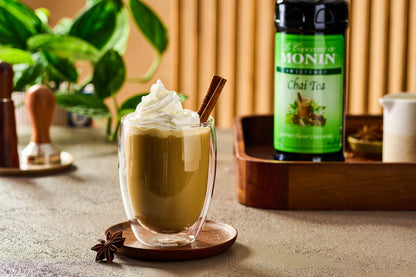 Monin Chai Tea Concentrate beside a creamy chai latte topped with whipped cream and a cinnamon stick, featuring warm chai spices.