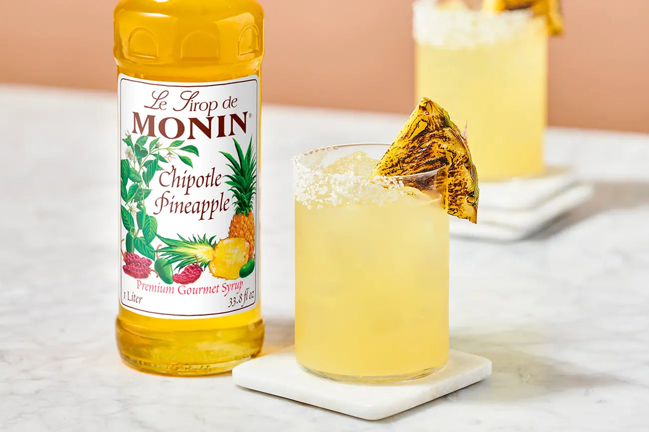 Monin Chipotle Pineapple Syrup beside a tropical pineapple cocktail with a salted rim and grilled pineapple garnish, featuring sweet and smoky flavour.