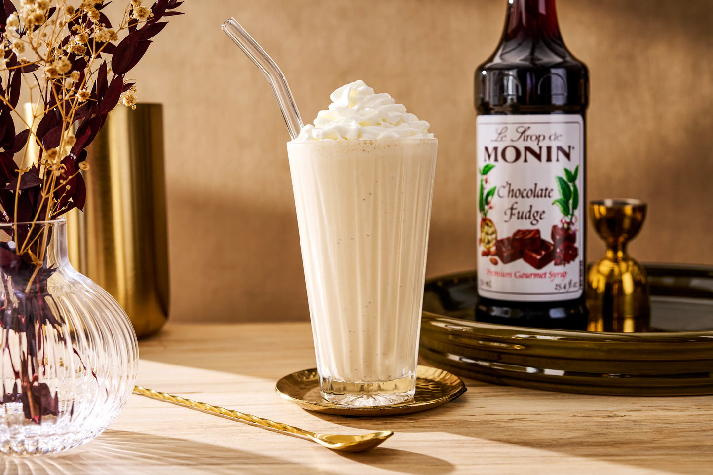 Monin Chocolate Fudge Syrup beside a creamy chocolate fudge milkshake topped with whipped cream, showcasing a rich dessert beverage.