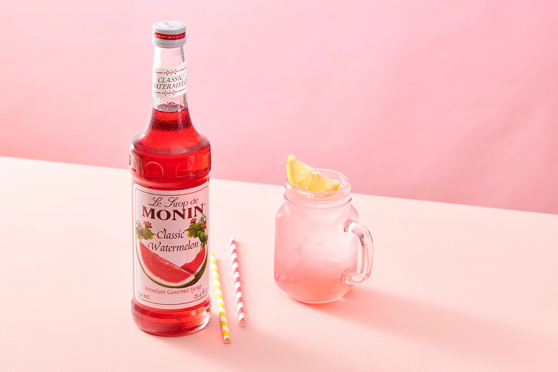 Monin Classic Watermelon Syrup beside a refreshing watermelon drink with ice and lemon garnish, showcasing fruity watermelon flavour.