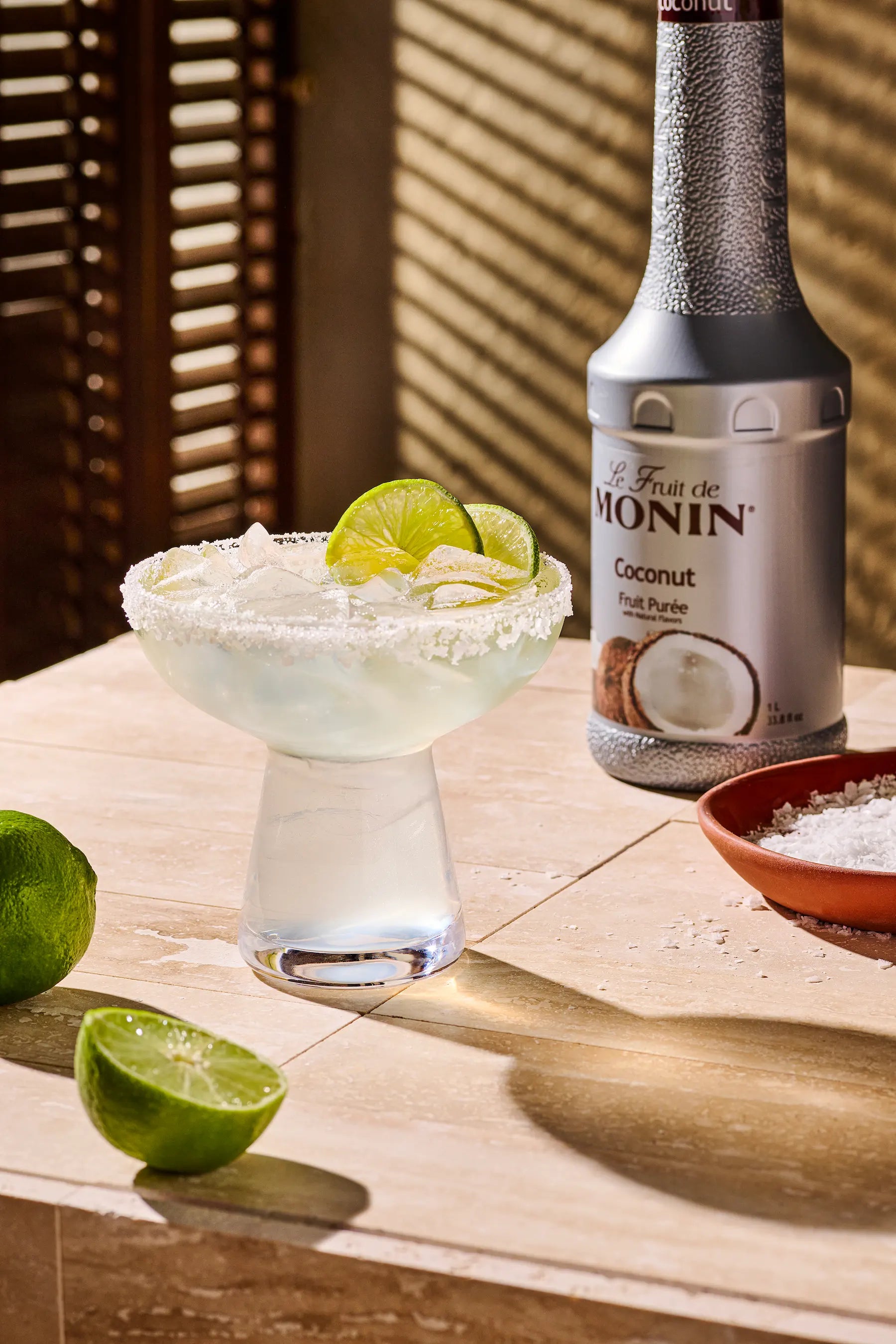 A bottle of Monin Coconut Puree next to a margarita glass filled with a pale cloudy liquid, garnished with lime slices, on a sunlit wooden table.