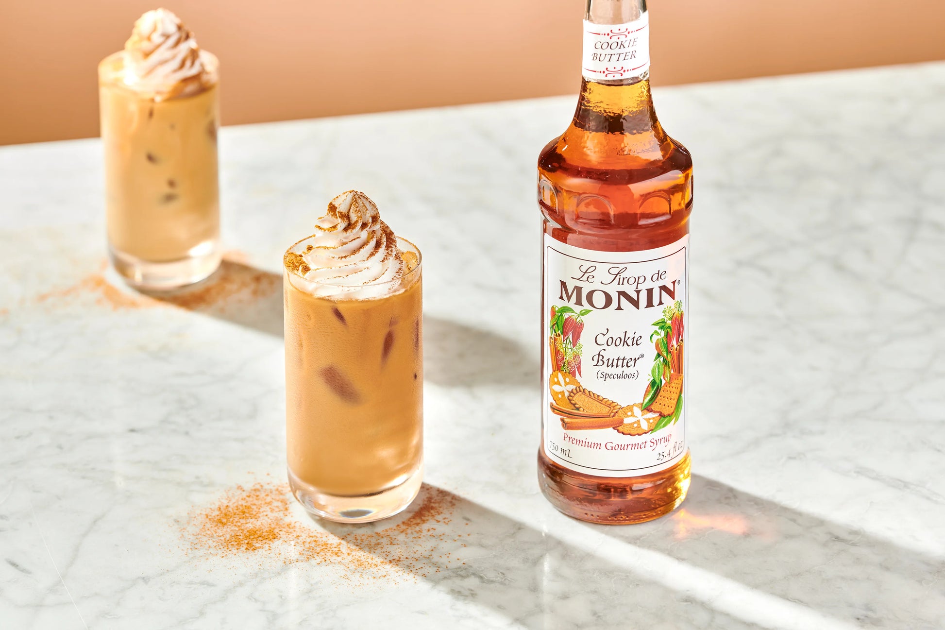 A bottle of Monin Cookie Butter Syrup stands on a white marble surface beside two iced coffee drinks topped with whipped cream, with a warm peach-toned background.