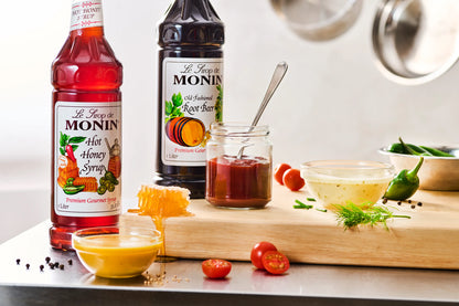 Monin Old Fashioned Root Beer Syrup with Hot Honey Syrup, sauces, tomatoes, peppers and honey on a kitchen counter.