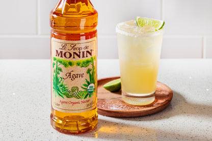 A bottle of Monin Agave organic nectar stands on a light countertop beside a margarita glass garnished with lime, on a round wooden coaster.