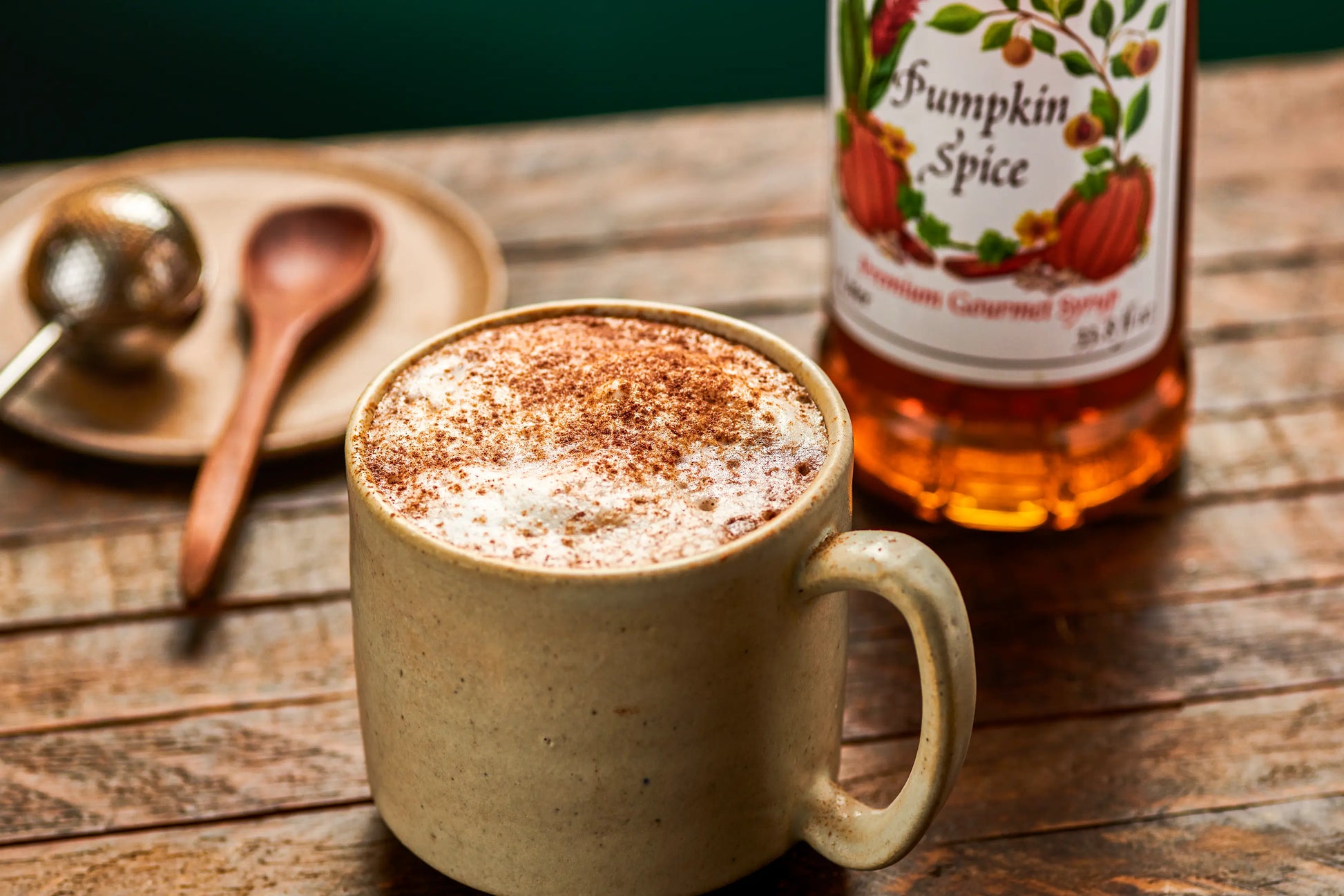 Monin Pumpkin Spice Syrup bottle beside a pumpkin spice latte topped with foamed milk and cinnamon on a rustic wooden table