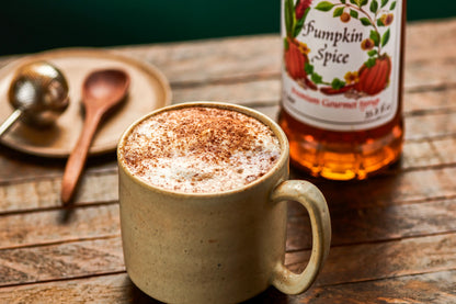 Monin Pumpkin Spice Syrup bottle beside a pumpkin spice latte topped with foamed milk and cinnamon on a rustic wooden table